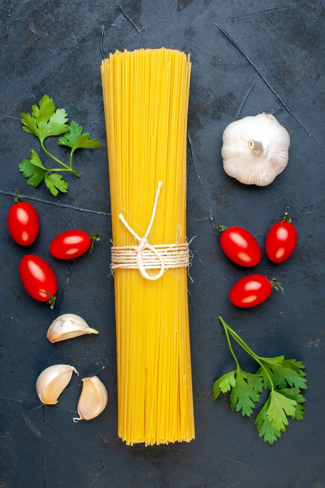 Draufsicht: ein Bündel Spaghetti, Cherrytomaten, Knoblauch und Petersilie auf schwarzem Hintergrund.